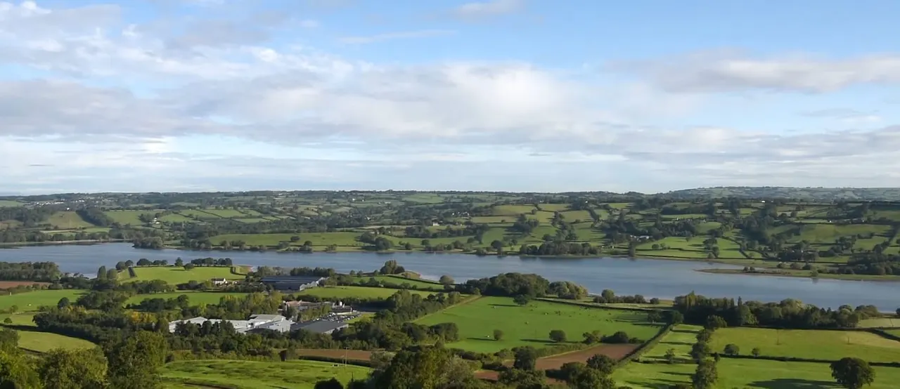 Aerial view of rolling countryside with green fields, two lakes, scattered trees, and farm buildings dotted across the landscape.