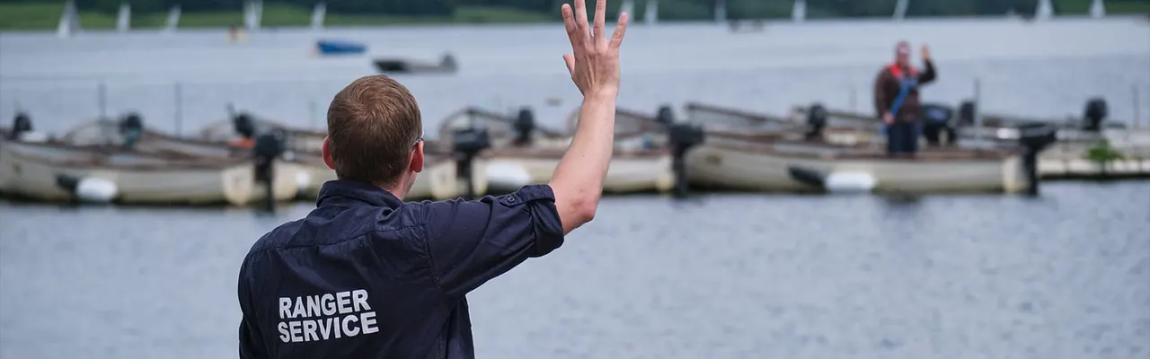 A ranger in uniform waves toward the water from the shore, with moored sailing boats visible in the background.
