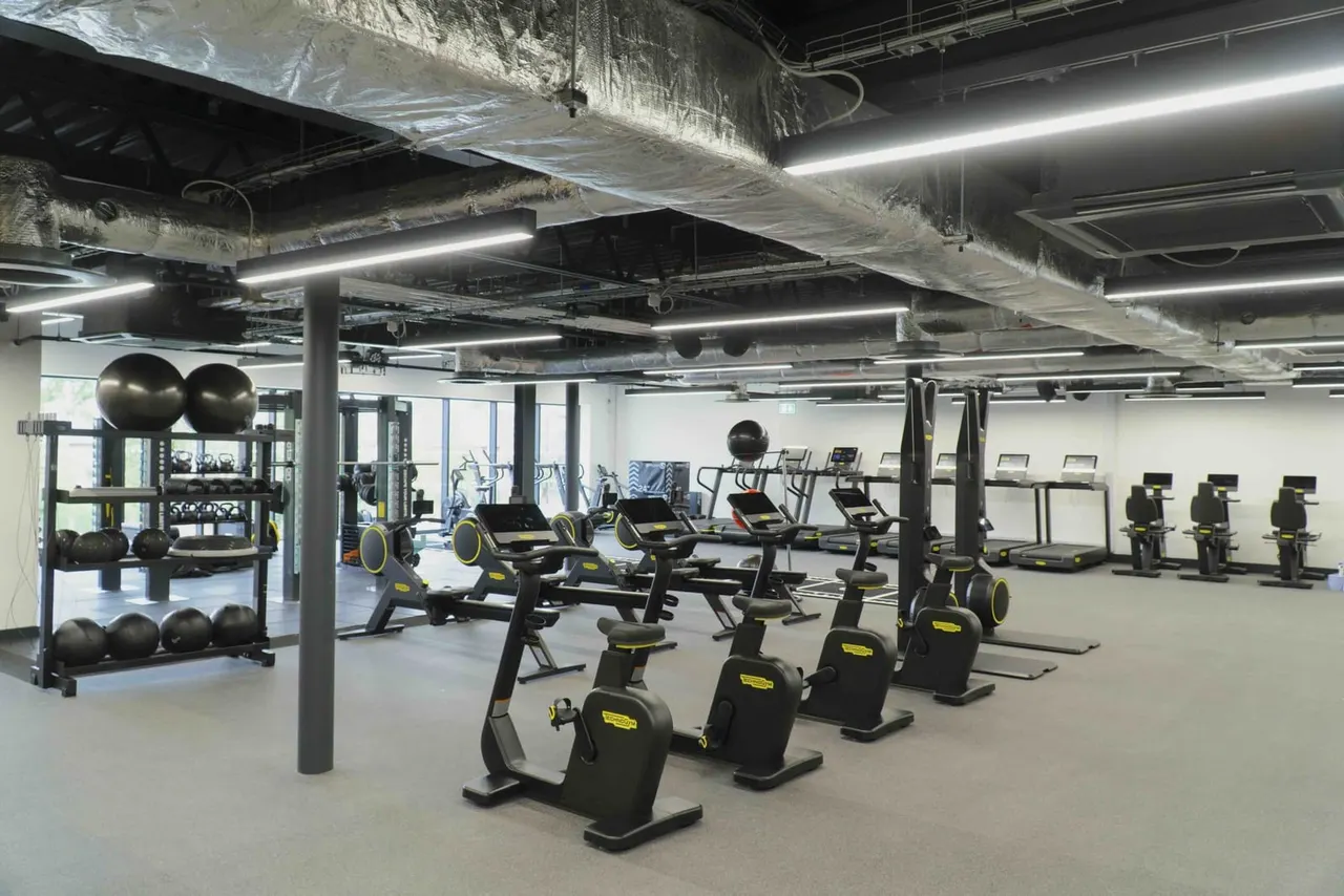 Spacious gym interior with rows of exercise bikes and stationary equipment, weights storage, and industrial ceiling.