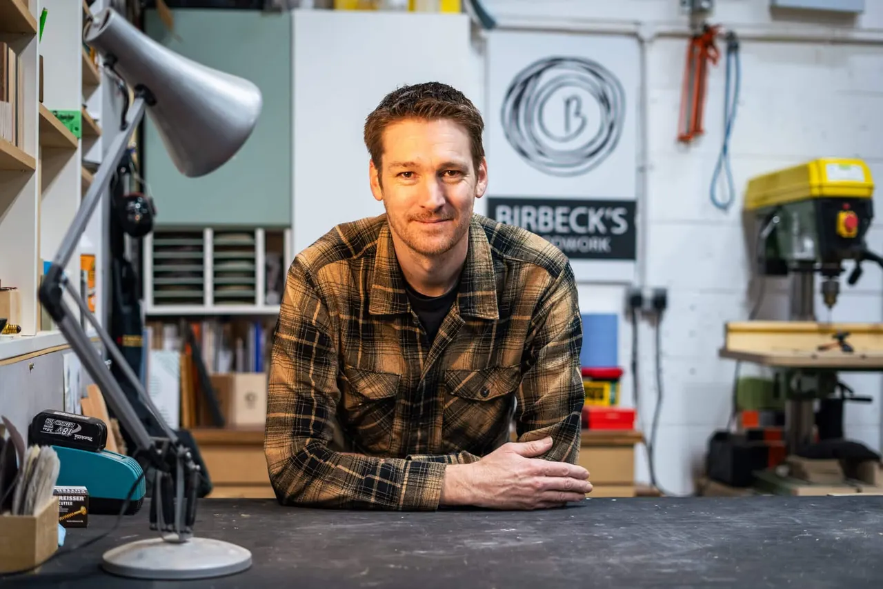 Man in checked shirt at workbench in workshop with tools and equipment on walls behind him.