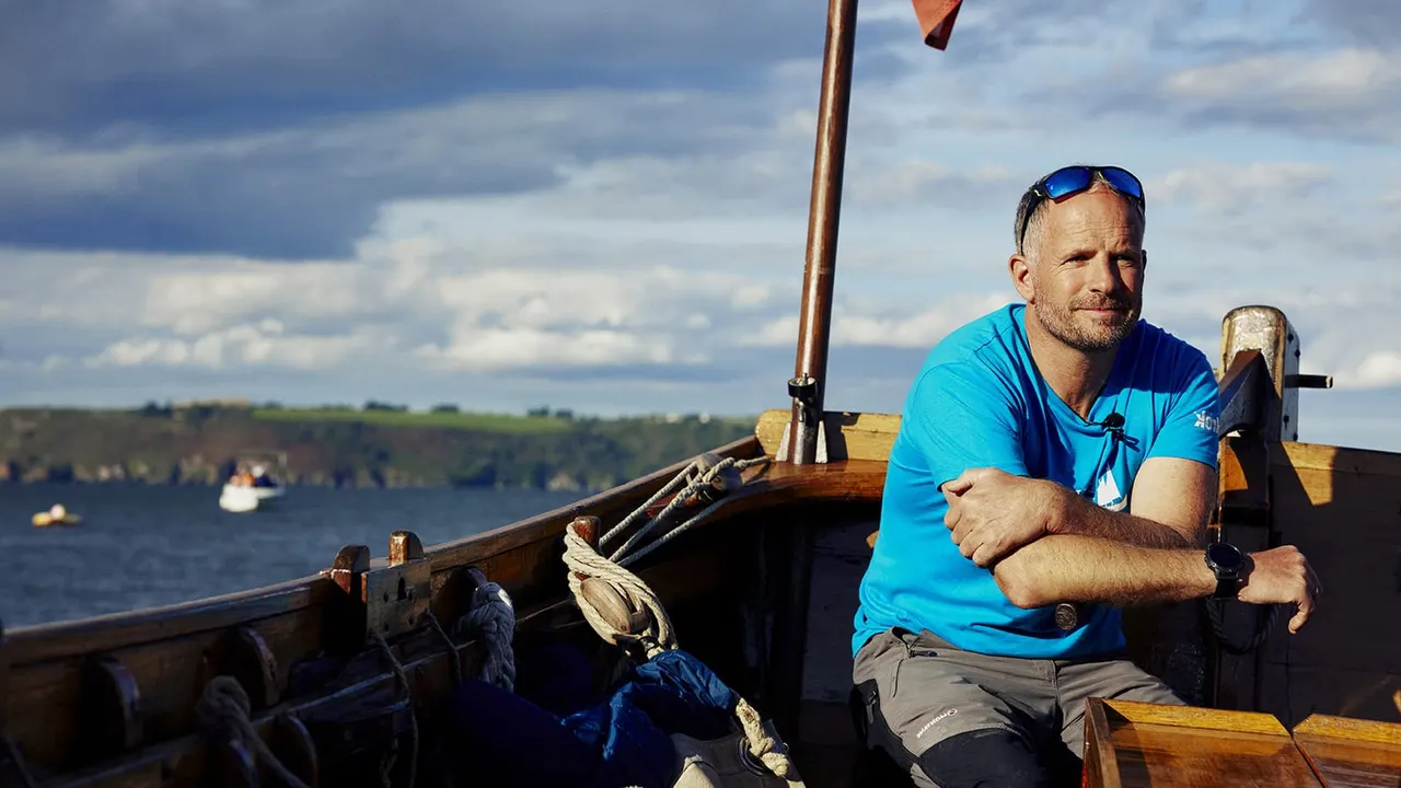 Man in blue shirt sitting aboard a wooden boat on water with wooded coastline in background.