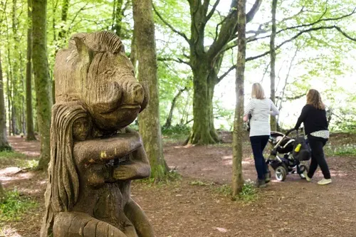 Wooden squirrel sculpture in woodland with two people and a pram on a forest path behind it.