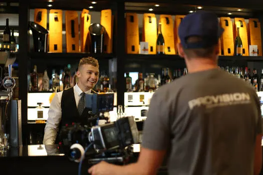 Bartender in waistcoat serving customer at illuminated bar with spirits bottles displayed behind.