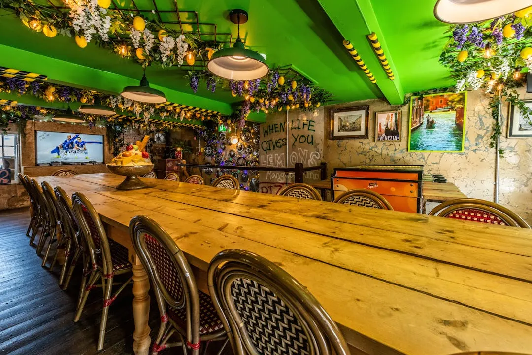 Long wooden table with chairs in a bright green-ceilinged dining space decorated with hanging flowers, bunting, and quirky wall art.