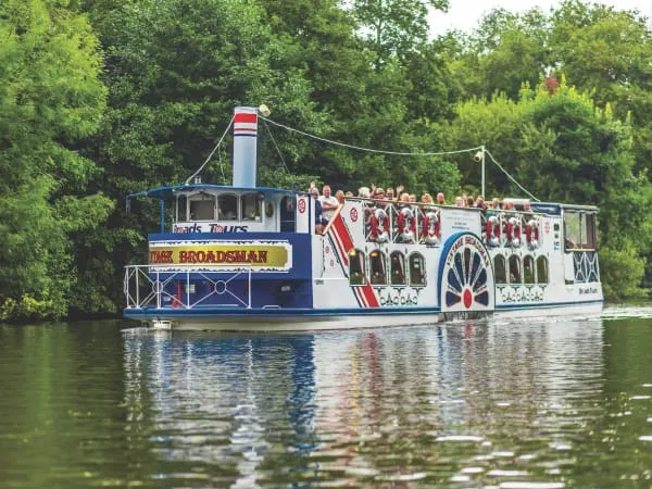 A paddle steamer riverboat with passengers aboard cruises along a tree-lined waterway.