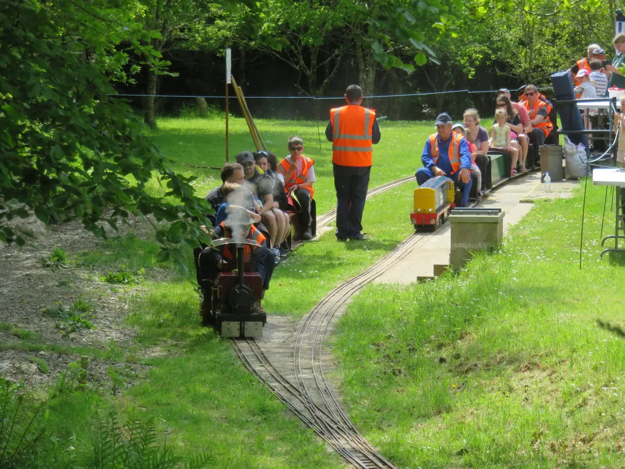 Children riding in miniature steam train carriages along narrow-gauge tracks through a grassy park with trees, supervised by staff in orange hi-visibility vests.