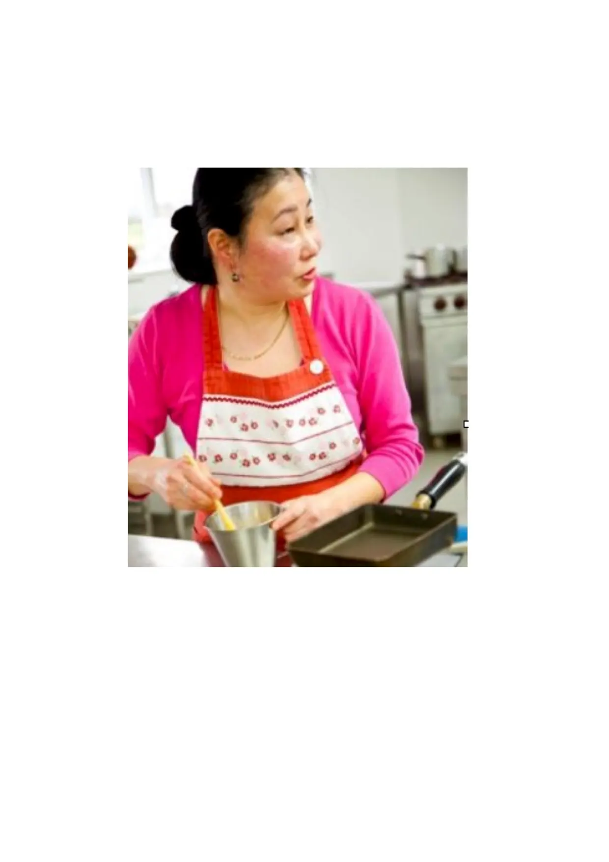 Woman in a pink cardigan and patterned apron preparing food in a kitchen during a cooking class.