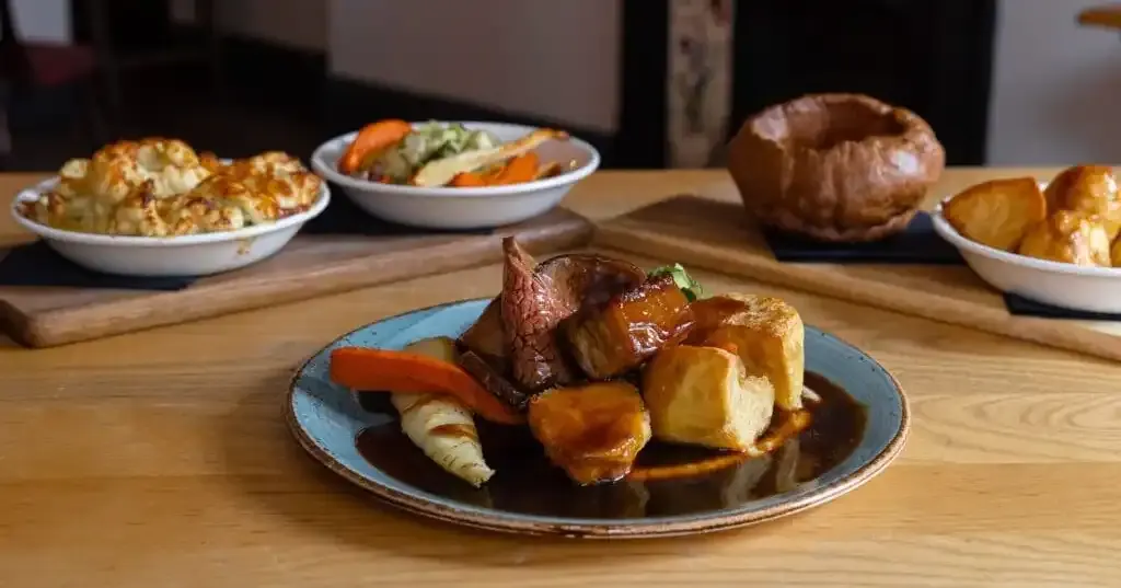 Roasted meat with potatoes and vegetables on a blue plate, surrounded by side dishes including salad, Yorkshire pudding and roast potatoes.