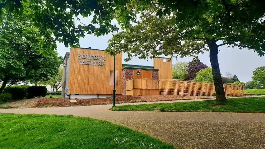 Modern wooden theatre building with green roof surrounded by mature trees and landscaped grounds.
