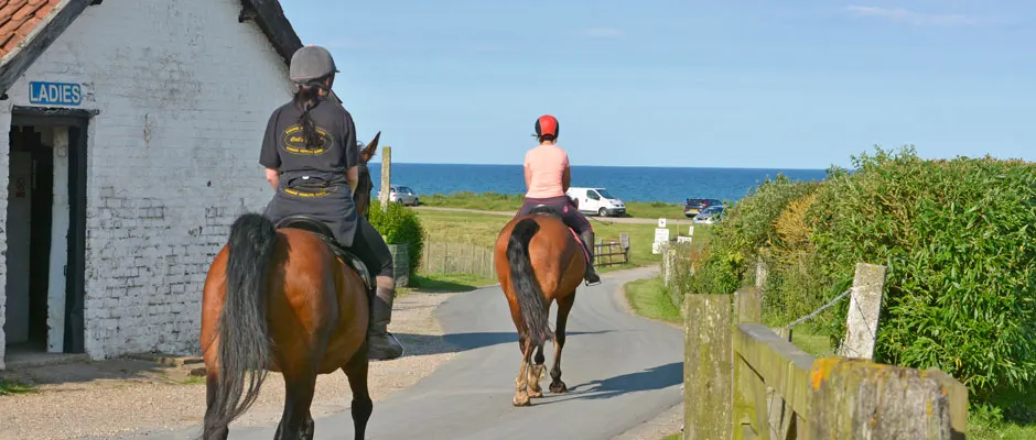 Two riders on horses on a coastal path with beach and sea in the background.