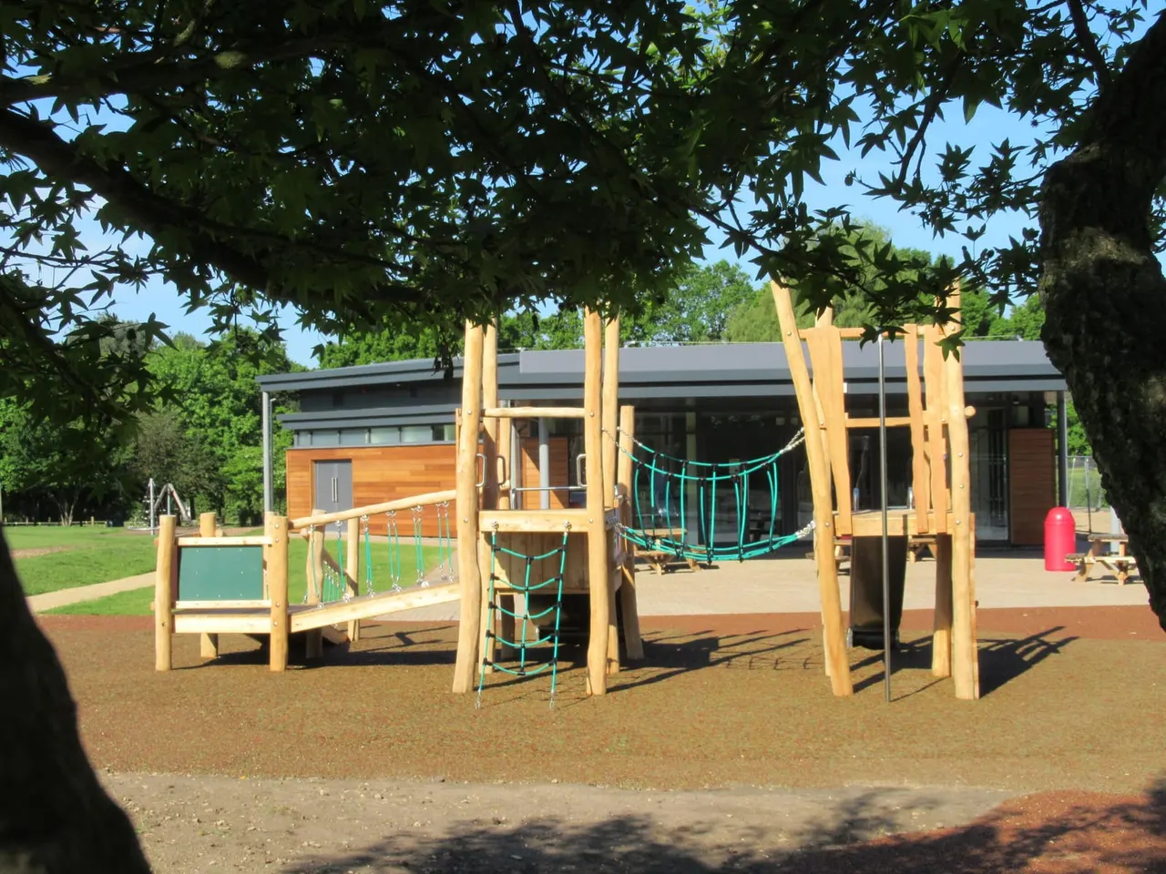 Wooden climbing frame with rope nets and platforms on a bark-covered playground, with a modern building and green space beyond.