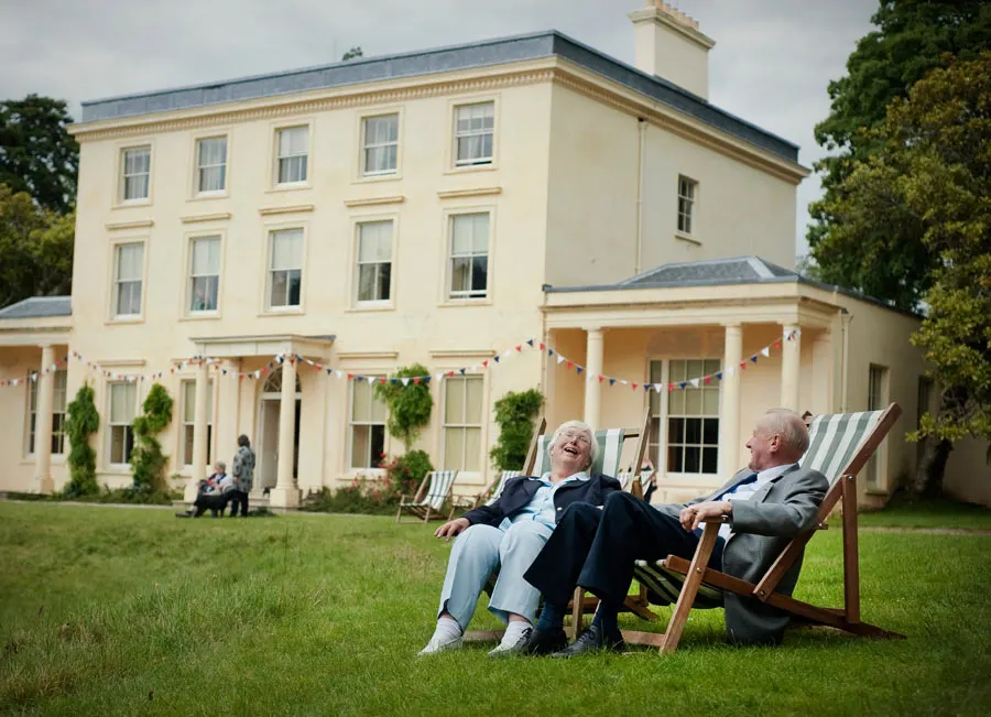 Two older adults relaxing in deckchairs on a lawn in front of a cream-coloured Georgian mansion with bunting.