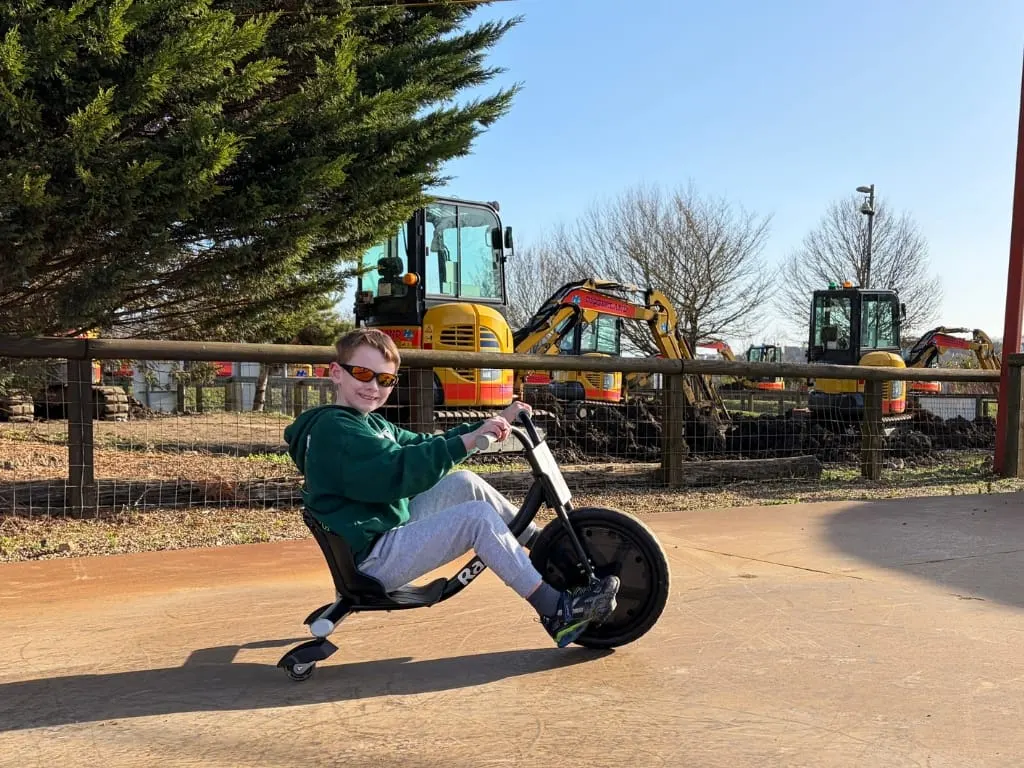 Child riding a drift trike on a sandy path with diggers and construction vehicles behind a fence.