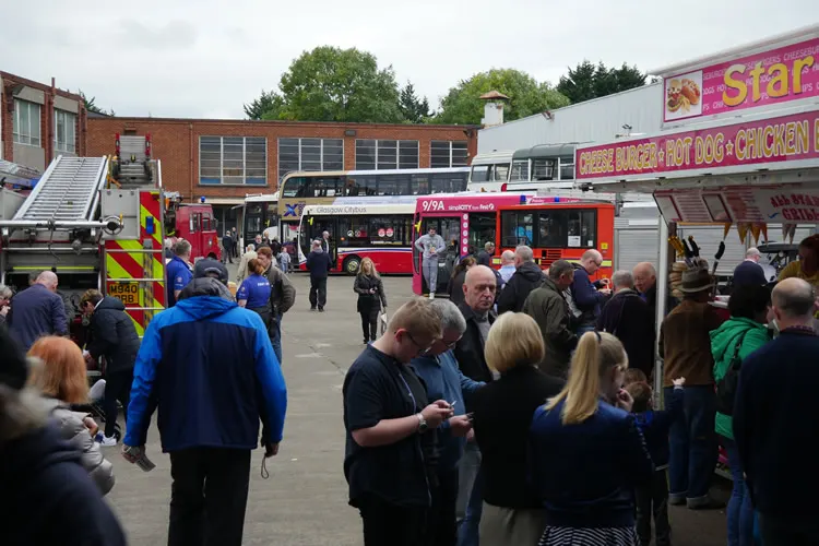Crowds of visitors queue at food vendor vehicles in a vintage bus garage yard, with historic buses and fire engines on display.