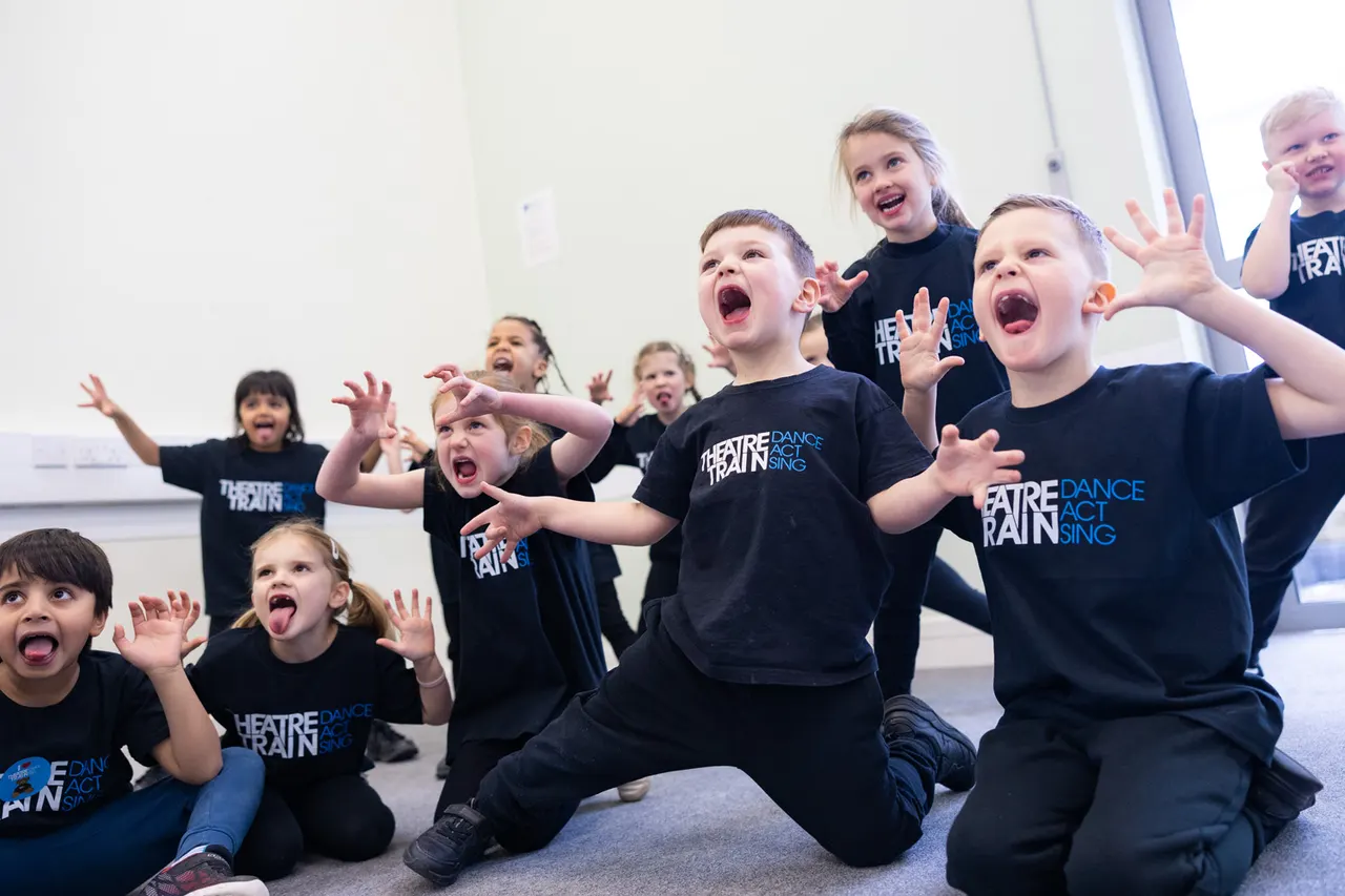 Group of children in black theatre training t-shirts performing an enthusiastic, animated routine indoors with exaggerated expressions and gestures.