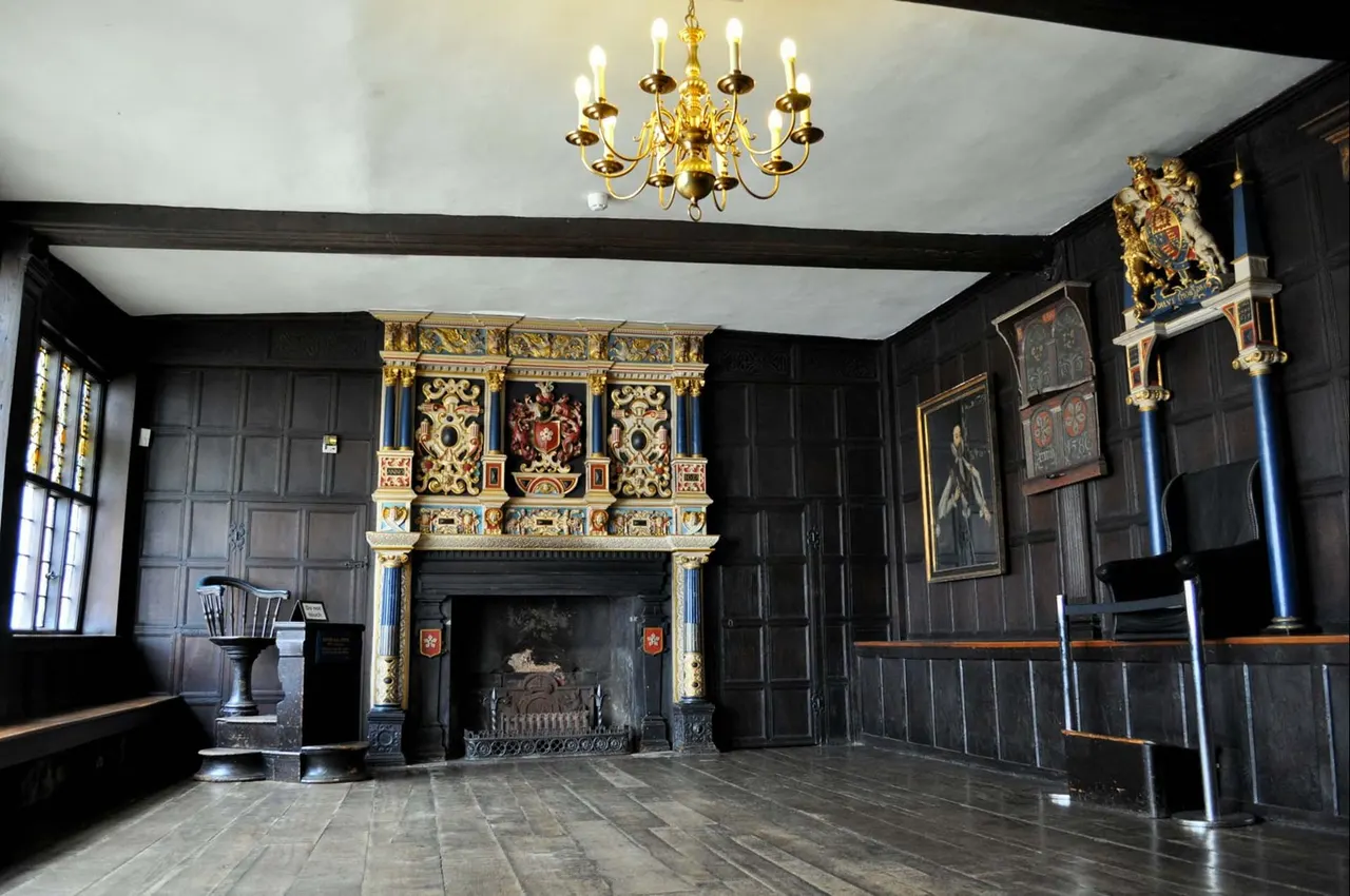 Historic black-panelled hall with ornate gilt fireplace, gold chandelier, and heraldic paintings on walls.
