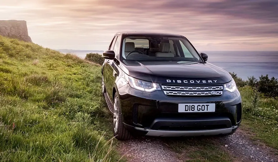 Dark Discovery vehicle parked on a grassy hillside overlooking a sea coast at sunset.