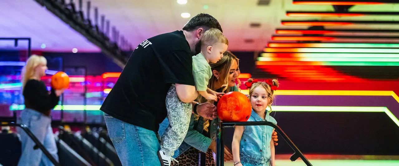 An adult and three children hold orange bowling balls at a brightly lit bowling alley with neon signage.