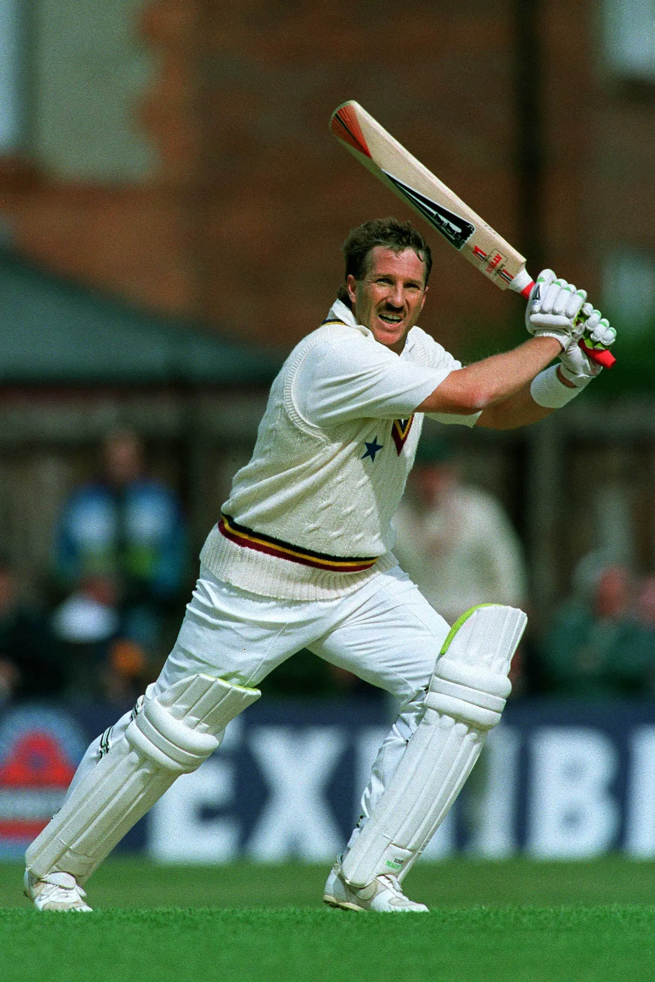 A male cricketer in white uniform and pads plays a shot with bat raised during a match.