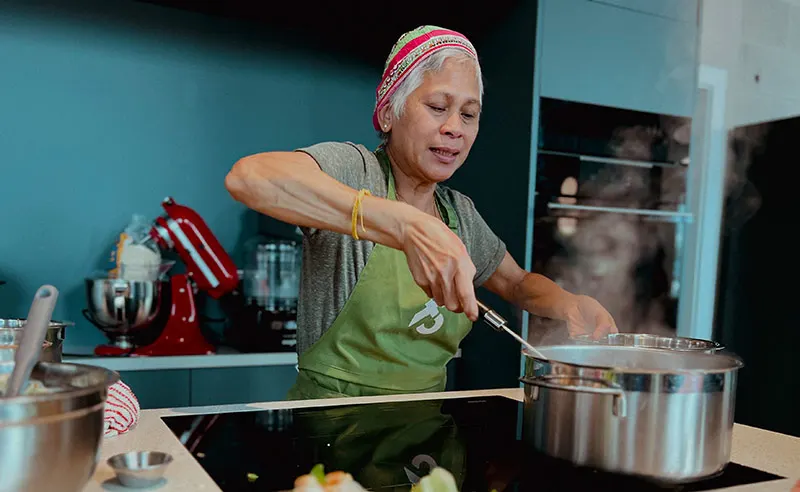 Woman in green apron and red headwrap stirring a pot during a cooking class in a turquoise kitchen.