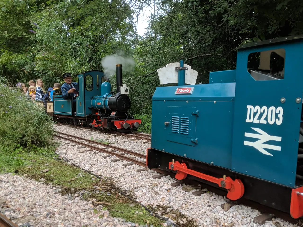 Passengers board a small steam locomotive on a narrow-gauge railway track surrounded by dense green foliage.