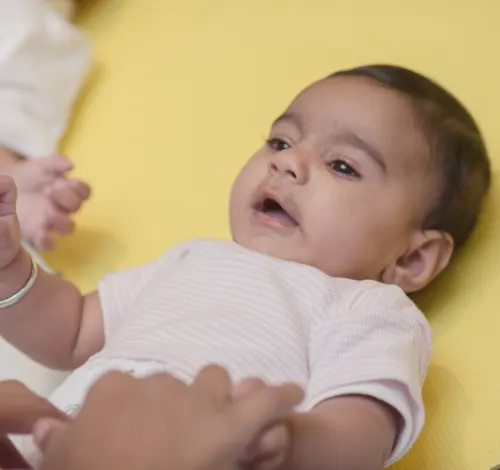 Baby lying on yellow background in white clothing, being held by an adult's hands.