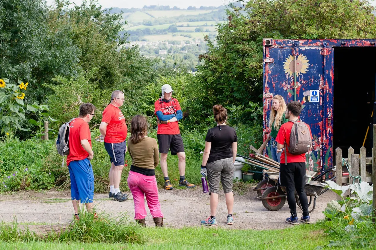 Group of visitors and staff in red shirts gathered outdoors at a rural farm location with rolling countryside in the background.