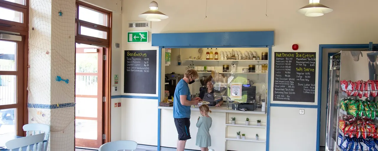 Adult and child at counter ordering food in a bright café with blue-framed servery, menu boards, and snack shelves visible.