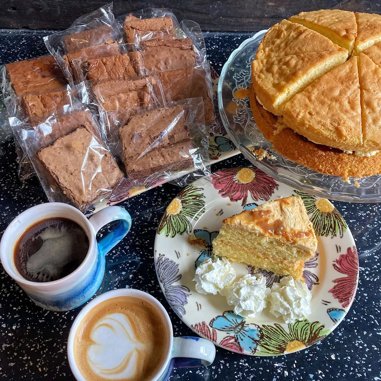 Selection of baked goods including brownies, sponge cake and pastries served with two coffees on a speckled dark surface.
