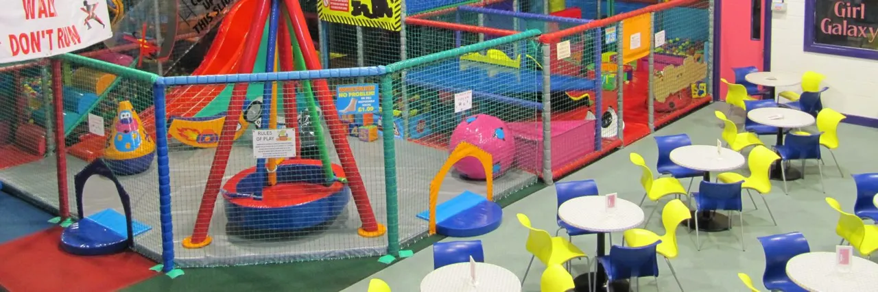 Colourful indoor play area with enclosed soft play structures, trampolines and tunnels behind netting, with bright yellow and blue chairs around white tables in foreground.