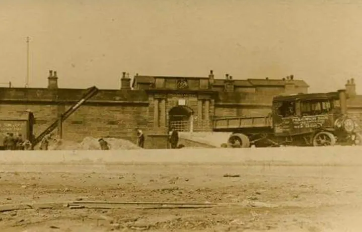Vintage sepia photograph of a stone fortification building with a vehicle and figures outside.