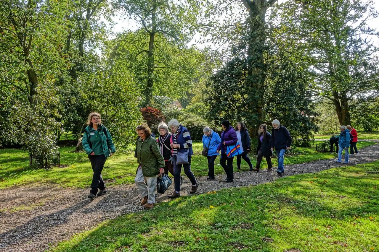 A guided group walk along a tree-lined gravel path through leafy woodland, with visitors of various ages enjoying the outdoors.