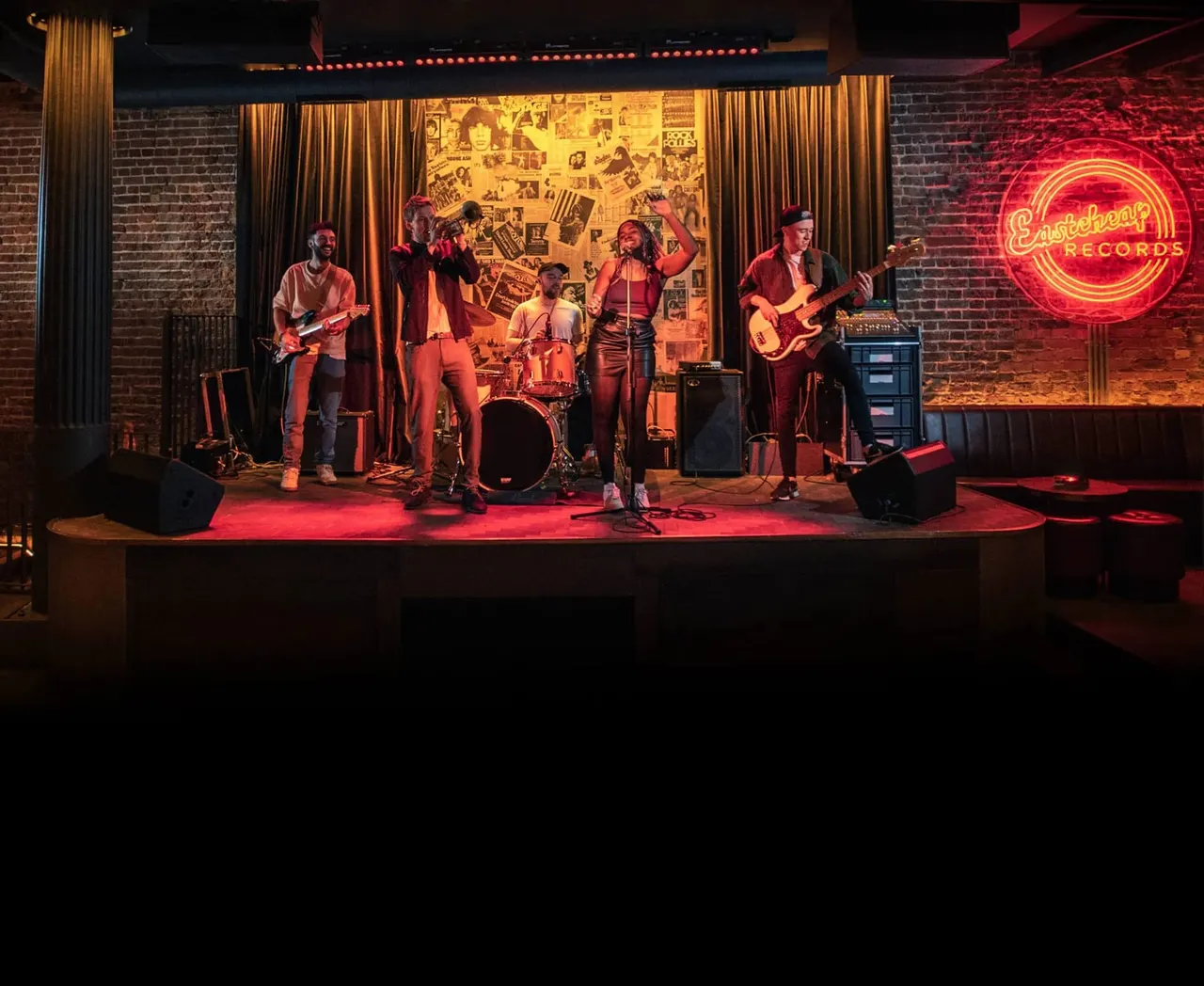 Live rock band performing on stage with red lighting at an indoor music venue with brick walls.