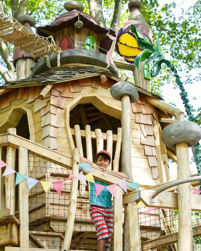 A child stands on a wooden platform decorated with bunting, part of a whimsical treehouse structure with carved details and colourful painted windows.