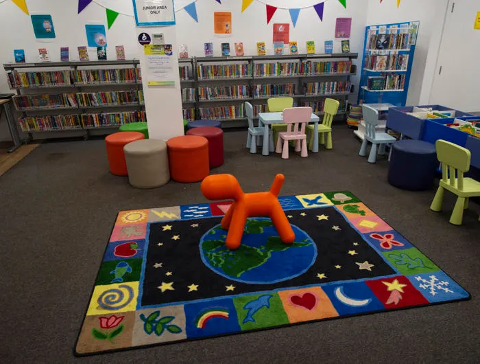 Children's library area with colourful play mat, reading cushions, small furniture, and bookshelves lining the walls.