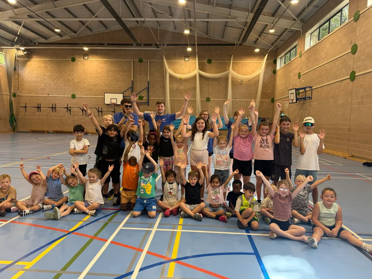 Large group of children of mixed ages posing happily in a sports hall with colourful floor markings and basketball hoops.