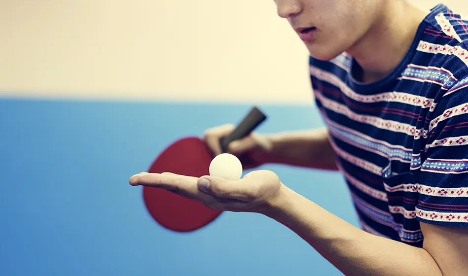 A young person holds a white ping pong ball on their palm whilst holding a red paddle, ready to play table tennis.