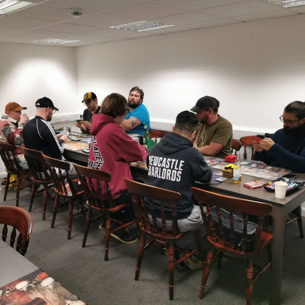 Group of people seated around tables in an indoor room, engaged in a tabletop gaming activity.