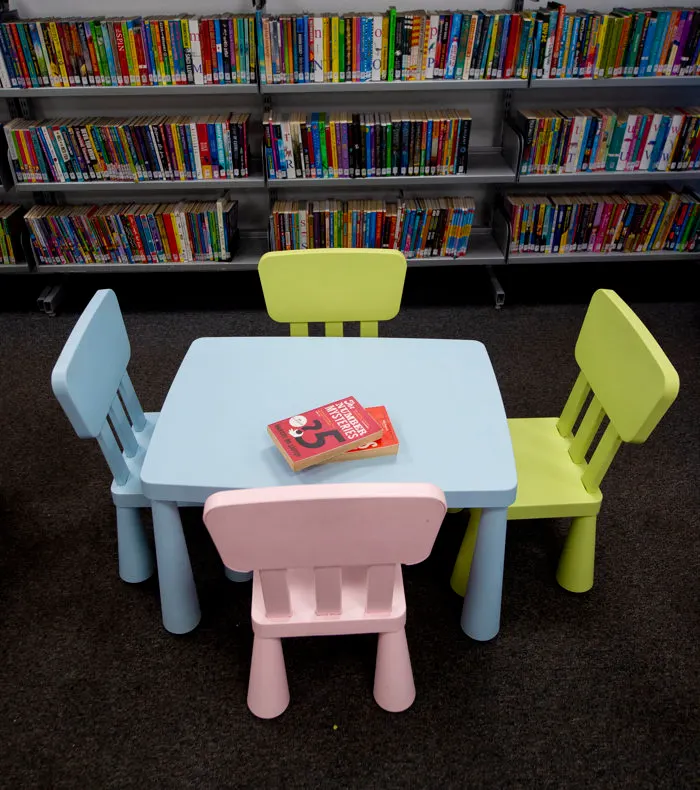 Child-sized table and chairs in a library, with coloured children's books on shelves behind.