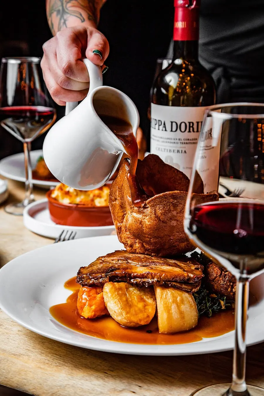 Person pouring sauce over roasted meat and potatoes at a fine dining table with wine bottles and glasses.