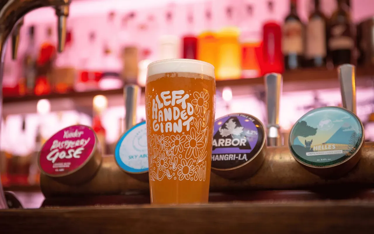 Glass of pale ale on bar counter with pump clips and pink-lit interior.