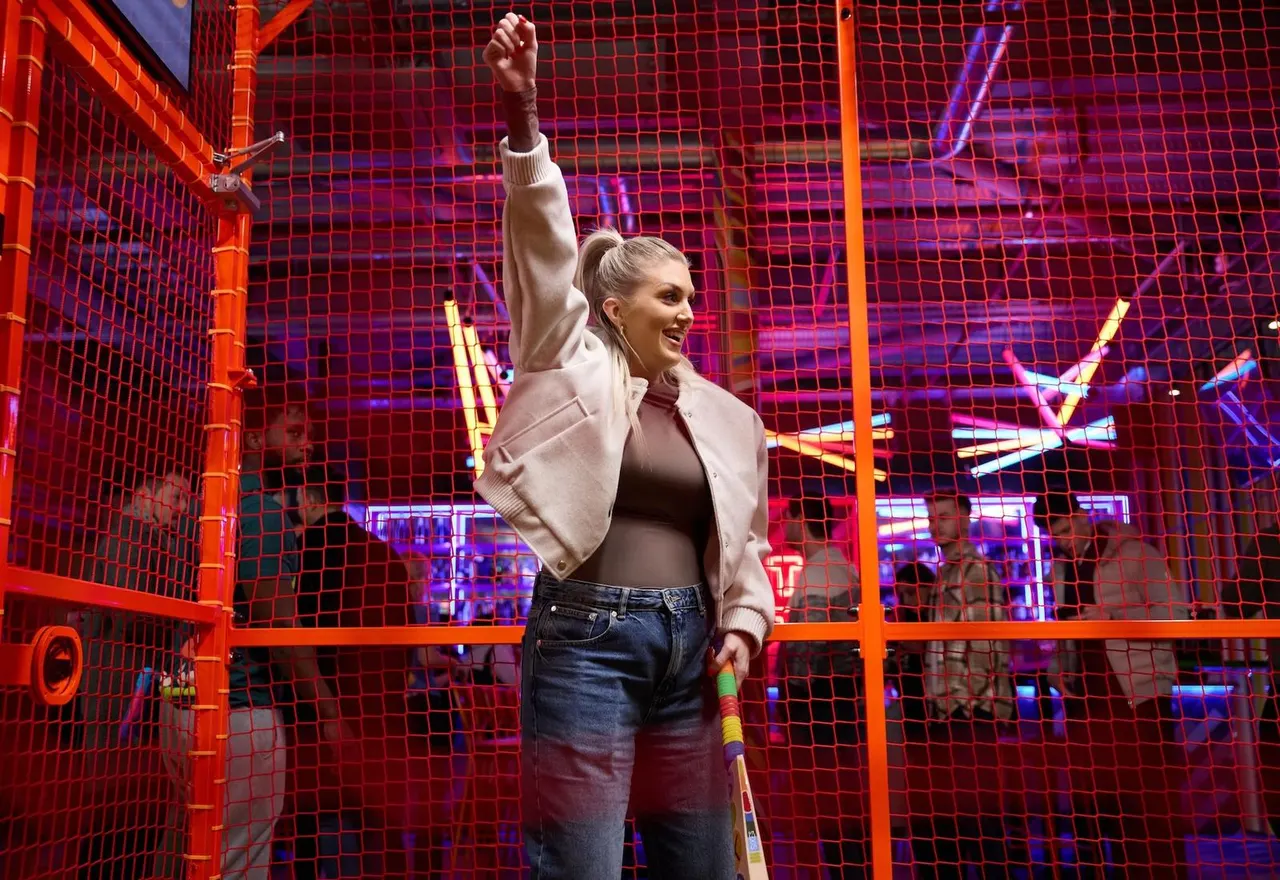 A woman celebrates in a neon-lit indoor cricket cage with a raised fist, holding a bat.