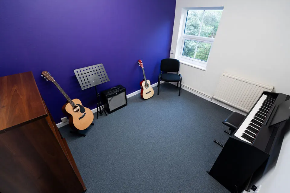 Music lesson room with acoustic guitars, keyboard, amplifier and music stand against purple wall.