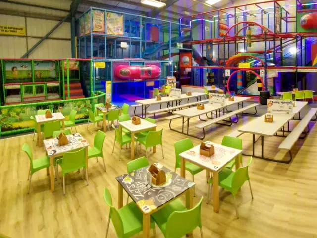 Indoor play centre with green tables and chairs in foreground, colourful climbing structures and ball pits visible throughout the large warehouse space.