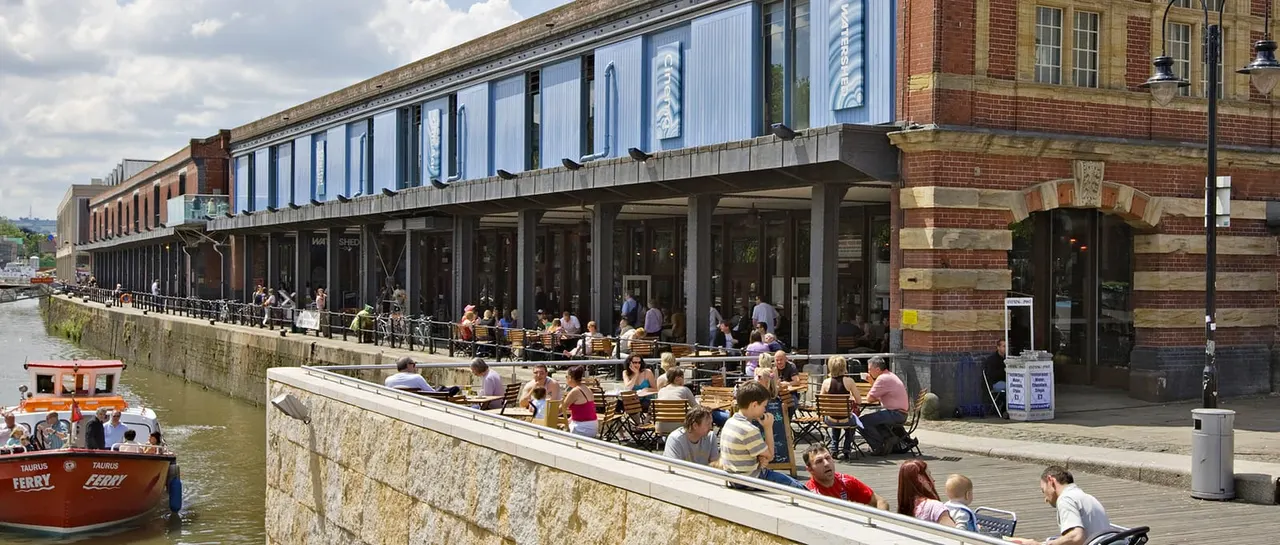 Waterside dining and leisure venue with visitors seated at outdoor tables along a canal, brick buildings behind.