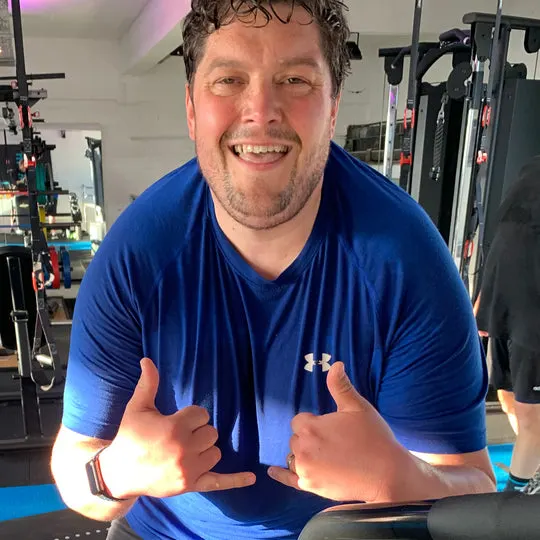 Man giving double thumbs up in a gym with exercise equipment visible in the background.