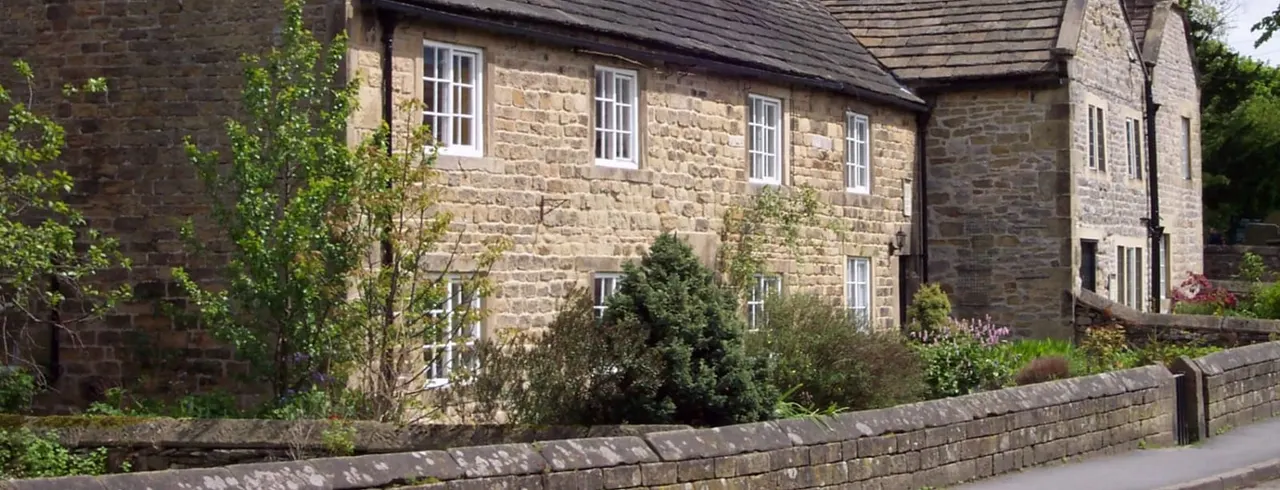 Stone-built cottage with multiple windows and a slate roof, surrounded by shrubs and climbing ivy.