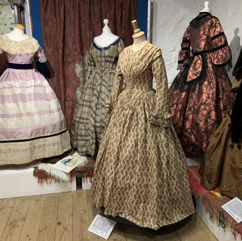 Museum display of Victorian and Edwardian era dresses on mannequins, including striped, checked and patterned fabrics in muted colours.