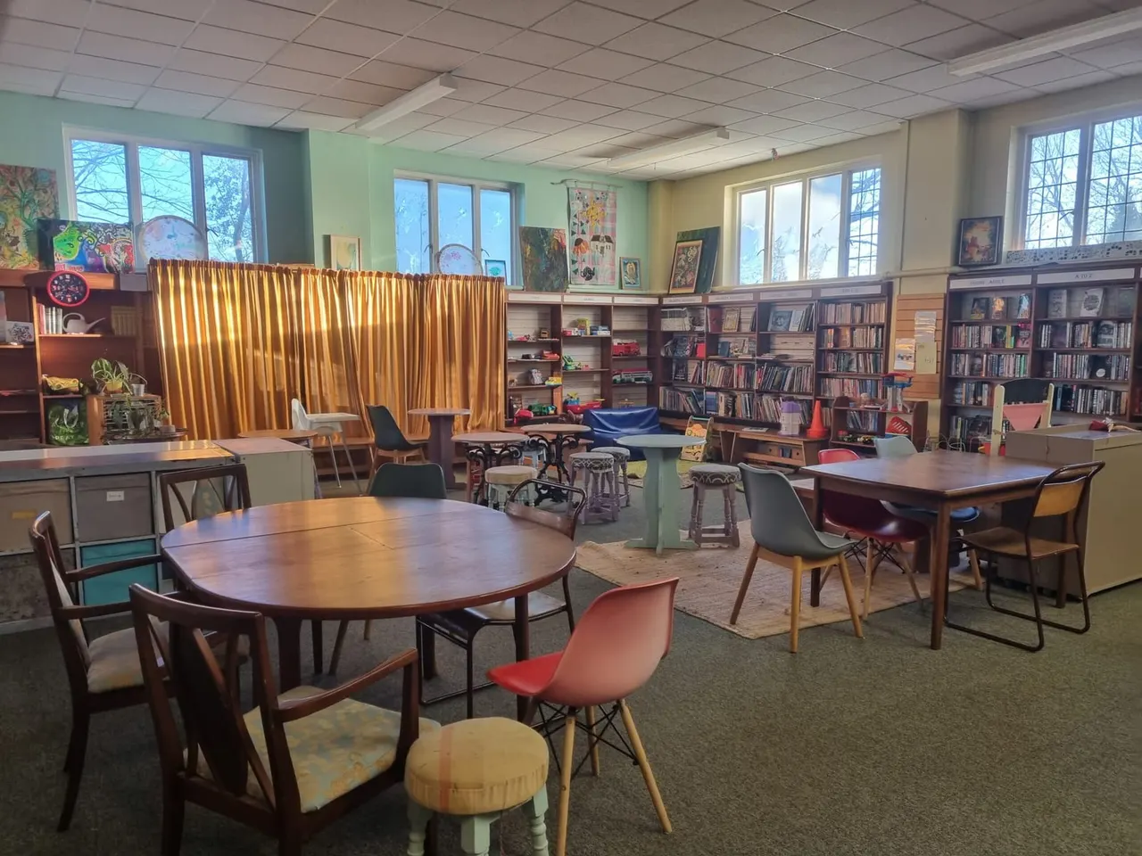 Light-filled community space with round wooden tables, mixed seating, bookshelves, artwork displays, and gold curtains beside windows.