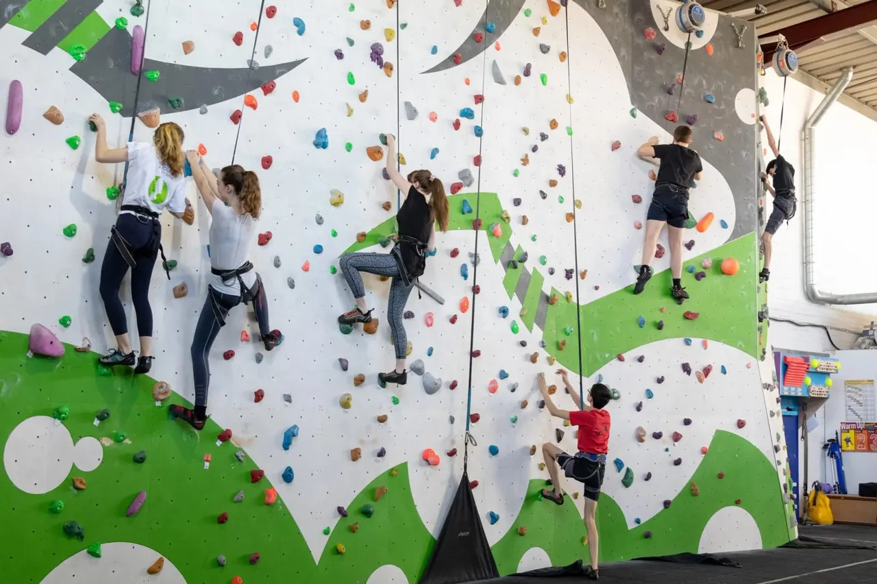 Multiple climbers of varying ages ascending coloured holds on large indoor climbing walls with safety ropes.